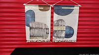 Pair of printed fabric wall hangings with neutral abstract shapes in brown, beige, blue, and gold, hanging side by side against a red background.