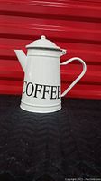 Front side view of the white enamelled metal coffee pot against red background, showing 'COFFEE' lettering, lid and handle.