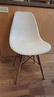 Front view of white molded plastic chair with pale wooden legs and black metal supports on hardwood floor.