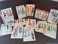 Wide view of eleven vintage 1960s sewing patterns from McCalls, Simplicity, Butterick arranged standing and lying flat against a purple background. Includes colorful fashion illustrations on paper envelopes.