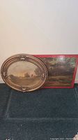 Two framed items against wall and floor, showing an oval ornate wood frame with rustic photo and rectangular red bordered wood frame with countryside photo.