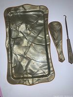 Full view of gold-toned dresser tray with textured marbled surface pattern and irregular wavy edge alongside matching shoe horn and hook tool, all metal with slight patina.