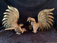 Front angle view of both brass rooster sculptures showing detailed wing and tail feathers.