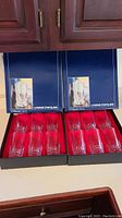 Two original navy blue boxes, each containing 6 clear crystal glasses with intricate cut pattern near the base, arranged inside with red interior lining.