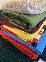 Photo showing stacked fabric cuts including knit tweed, wide green corduroy, silky suiting fabrics in burgundy, yellow, and multicolor striped textile, plus blue and red fabrics.