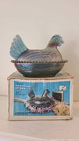 Iridescent carnival glass hen on nest sitting on original box, showing front detail and box art