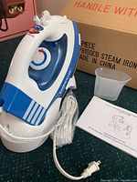 Oreck JP8100CB cord-free steam iron with original cardboard box, measuring cup, power cord wrapped and manual booklet showing the iron and box label.
