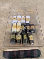 Clear plastic container holding 13 oversize spools of overlock thread in white, beige, green, black, and dark colors, sewing shears placed on top, and plaid sleeve ironing form beneath