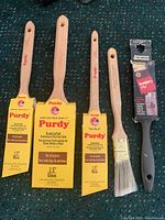 Five new paintbrushes laid out on green carpet: three Purdy brushes with yellow labels, a white bristle brush, and one black-handled angled brush by American Worksaver.