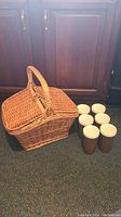 Wicker picnic basket and six brown and white insulated tumblers arranged on floor in front of wooden cabinets.