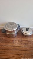 Two Lustre Craft stainless steel pots side by side on wood floor: Dutch oven with dome lid and black handles, smaller pot with lid.