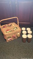 Tall wicker picnic basket with integrated handle decorated with 3D vegetable appliques on all visible sides, placed on carpet next to six vintage brown insulated tumblers with white interiors.