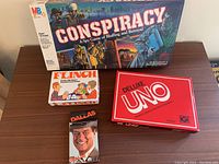 Group photo of four vintage board and card games: Conspiracy board game, Deluxe UNO, Flinch, and Dallas playing cards