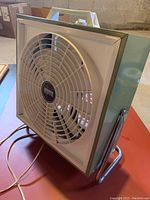 Side view of vintage Montgomery Ward portable table top fan showing olive green metal body, white front grille with fan blades inside, white handle on top, and cord extending.