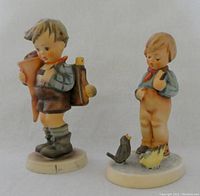 Front view of the two Hummel figurines side by side showing the boy with school cone and satchel and the boy looking at birds.