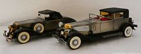 Photo showing both metal diecast cars side by side. The silver 1934 Duesenberg convertible lighter and the black 1931 Rolls Royce radio with hardtop roof and whitewall tires.