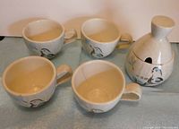 Four cream-colored ceramic coffee cups with handles and one ceramic sugar bowl with lid on blue surface, decorated with Bob White bird and floral pattern by Red Wing Pottery
