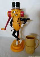 Front view of Mr. Peanut peanut butter machine standing with hand crank, black top hat, monocle, and a small tray. Yellow circular base, next to yellow plastic cup with peanut design.