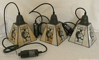 Three Kokopelli stained glass track lights with black ceramic bulb holders and attached cords. Each light has a square tapered shade with silhouettes of Kokopelli figures.