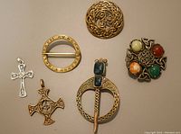 Top view of five Celtic-themed brooches and pendants including a knotwork circular brooch, a cross pendant, and a multi-stone brooch.