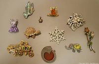 Top-down photo of a collection of 12 sparkly brooches and a small silver flower pendant featuring various designs including an elephant, turtle, snowflake, and carriage.