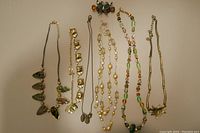 Photo of seven different green and bronze toned necklaces and one hair clip laid out flat on a table to show the entire collection and different styles.