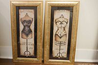 Pair of framed pictures leaning against a wall showing French corset illustrations with ornate gold frames.