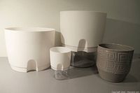 Three white self-watering pots with clear water reservoirs (one reservoir missing) and one taupe ceramic pot with a geometric pattern, shown side by side