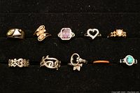 Top view of 8 rings arranged in two rows on black surface showing variety of designs, stones and sizes.