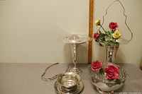 Two silver epergnes, large silver bowl with floral crosshatch insert, glass flower frog, silver flower holder, and several china roses arranged for display with a measuring ruler in the background.