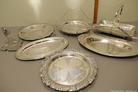 Full view of 7 serving platters arrayed showing various shapes, handles, and edge designs, including one leaf-shaped non-silver platter.