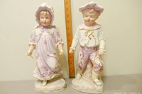 Full view of the ceramic boy and girl figurines standing side by side with a ruler showing their height at about 13 inches.