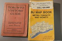 Two vintage map books placed on a grey surface. Left: orange-red 1939 Toronto Visitors' Guide. Right: yellow-cover 1986 Perly's BJ Map Book with business label on the front.