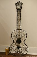 Full view of black wrought iron guitar sculpture with decorative scrolls and twisted edges, standing upright against a wall next to a can for scale.