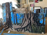 Wide view showing two mixed media artworks with large textured tree trunks, blue sky background, and cut log bases on carpeted floor.