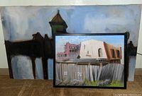 Two framed acrylic paintings displayed leaning against a wall, one larger with a train structure silhouette, one smaller with urban building scene.