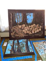 Both mixed media paintings showing stacks of wood rounds against a forest backdrop with blue sky, laid on carpet and partially framed.