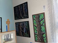 Three framed paintings hanging on a pale wall, showing two dark framed canvases stacked vertically and one framed canvas positioned next to them.