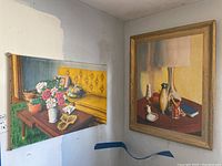Two acrylic paintings by Donald Gotz pictured side-by-side on a wall. One unframed painting leans on the wall showing a yellow tufted sofa with a table of flowers and objects. The other framed painting shows a lamp and other objects.