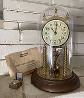 Front view of Kundo anniversary clock under glass dome with winding key, warranty envelope, and metal box quartz clock beside it.