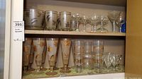 Shelved barware including pilsners with golden foliage, cocktail and stem glasses with silver foliage designs, and clear stemmed glasses.