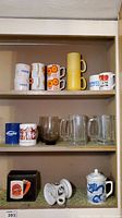 Shelf view showing assorted mugs including vintage orange flower design, yellow stacked mugs, personalized mugs, and clear glass beer mugs.