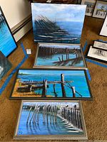 Four framed acrylic paintings laid on the floor showing coastal and dock scenes with wooden pilings in water, various sizes and compositions.