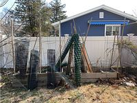Overview photo showing black metal wire plant cages leaning against wooden raised garden beds, bundled green plastic garden netting rolled up, and multiple wooden garden stakes standing upright.