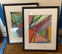 Two framed abstract art pieces side by side with black frames and white mats on a carpeted floor against a wall.