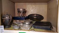 Photo of silver plated serving pieces including a teapot, chafing dish base, crumb butlers, trays, and glass casserole dishes on a wooden shelf.