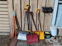 Five snow shovels leaning on a weathered wooden wall, two buckets of snow melt, two solar lights, and a bundle of orange plastic ground stakes next to a rectangular wood base with casters.
