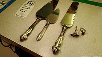 Overview of three sterling silver holloware handled cake/pie servers and two rings on beige surface