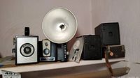 Front view of vintage box cameras including Ansco and Kodak Brownie Hawkeye with flash attachments, displayed on a shelf.