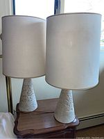 Image showing both vintage ceramic lamps on a wooden side table with large white fabric drum shades, lit by daylight through a window.